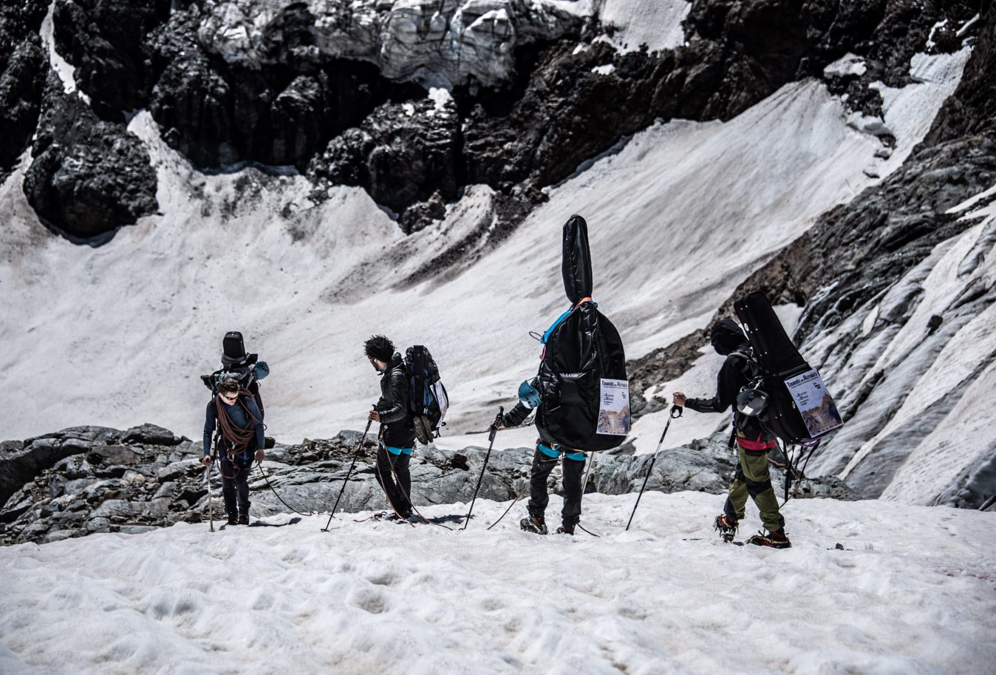 Quatre personnes encordées marchent sur la neige en haute montagne. Elles portent sur le dos, en plus de leur sac, des instruments de musique, dont une contrebasse.